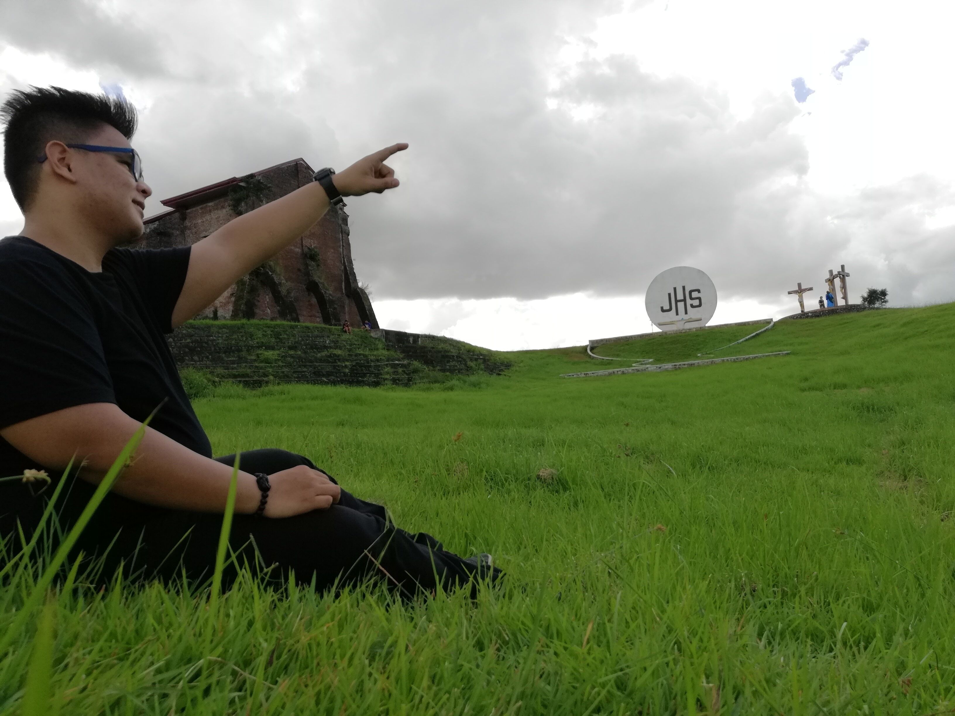A person sitting on grass pointing towards a hill with crosses and a large JHS sign.