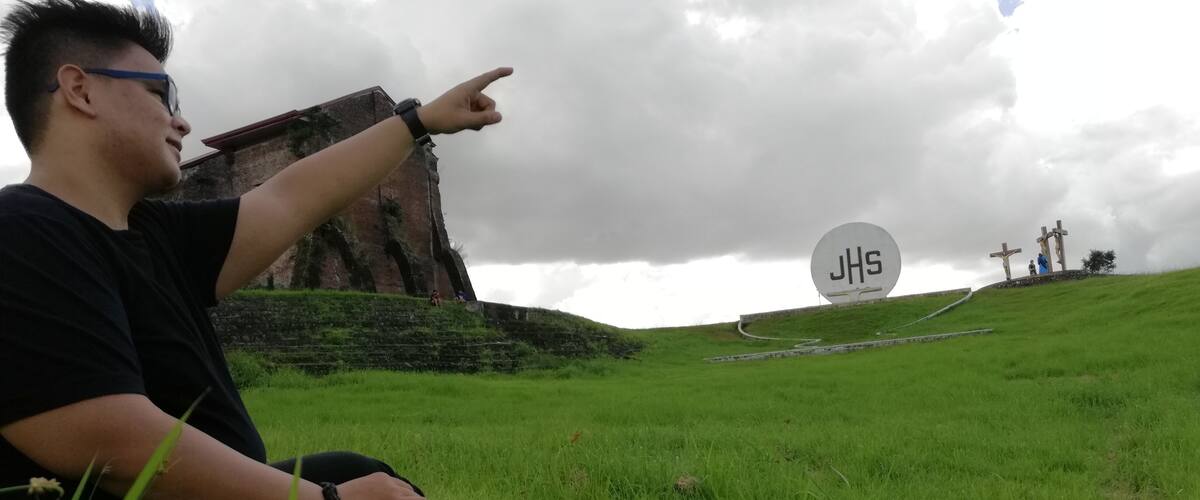 A person sitting on grass pointing towards a hill with crosses and a large JHS sign.