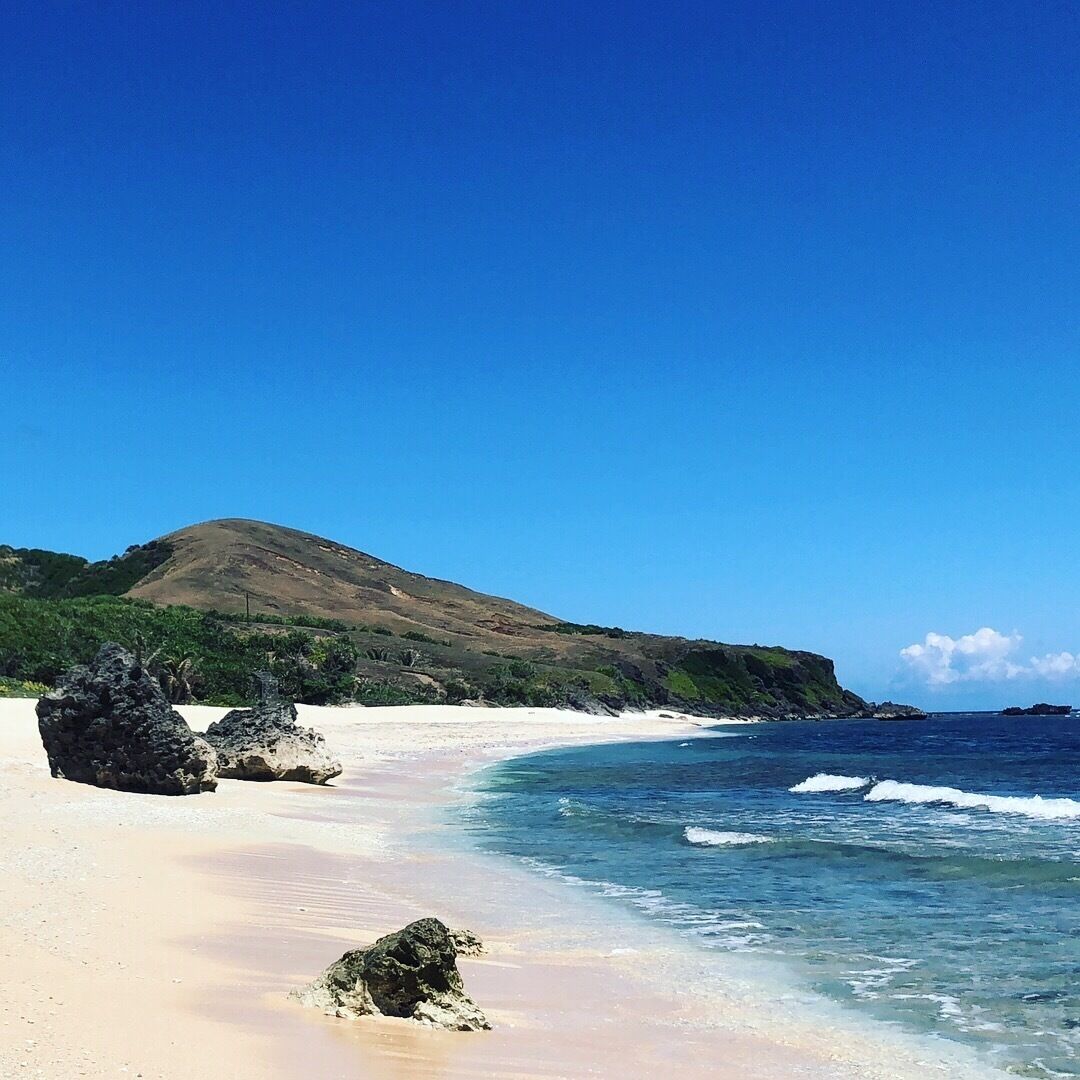 A beach with clear blue water, sandy shore, and a hill in the background.