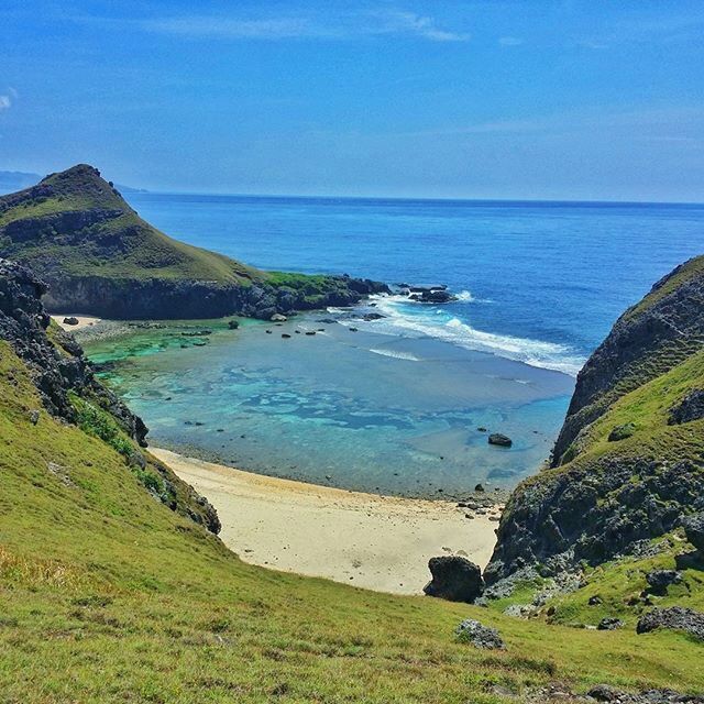 A coastal landscape with a clear blue sea, a sandy beach, and green hills.