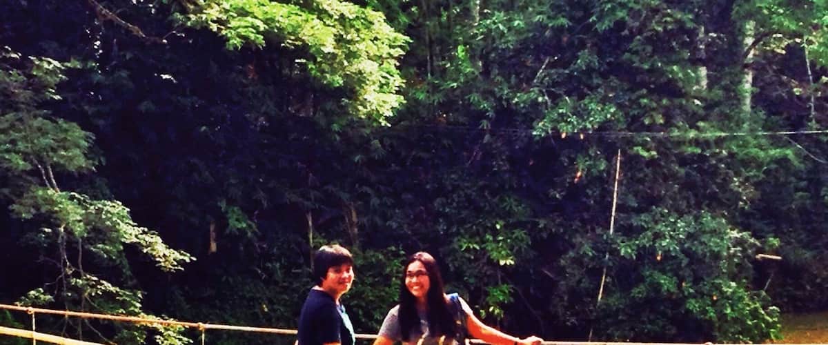 Two people standing on a bamboo bridge over a body of water, surrounded by dense green foliage.