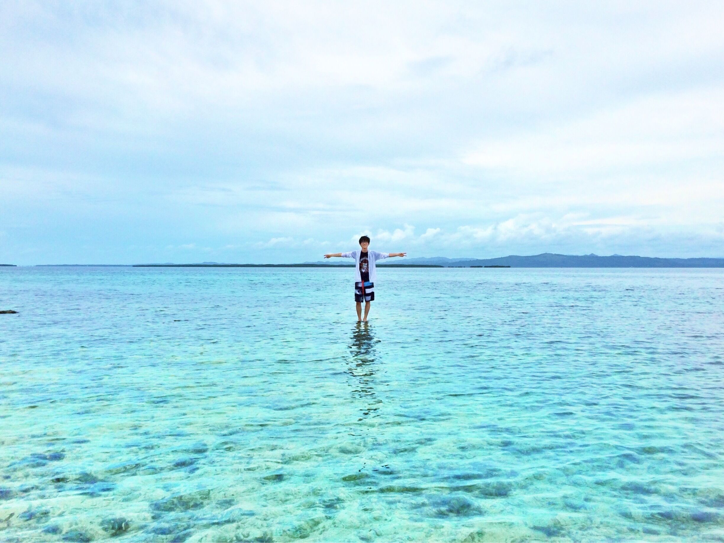 A person standing in clear shallow water with a distant shoreline and mountains.