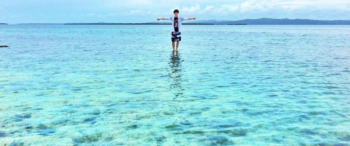 A person standing in clear shallow water with a distant shoreline and mountains.