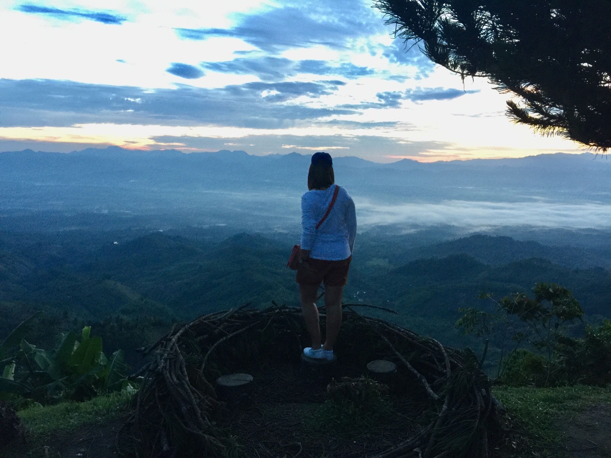 A person standing on a woven platform overlooking a mountainous landscape with a sunset in the background.