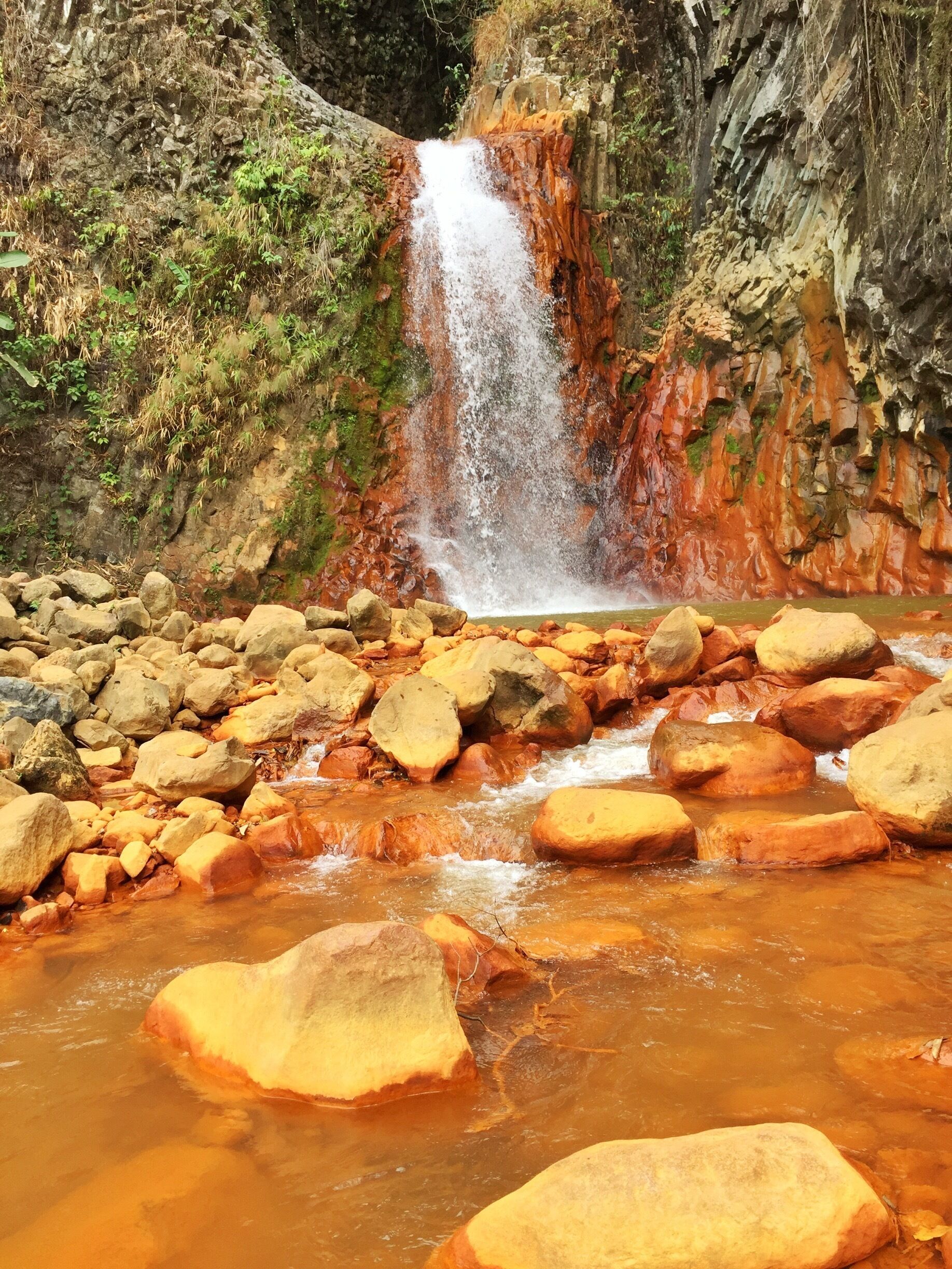 A waterfall cascading over reddish-brown rocks into a shallow stream with large boulders.