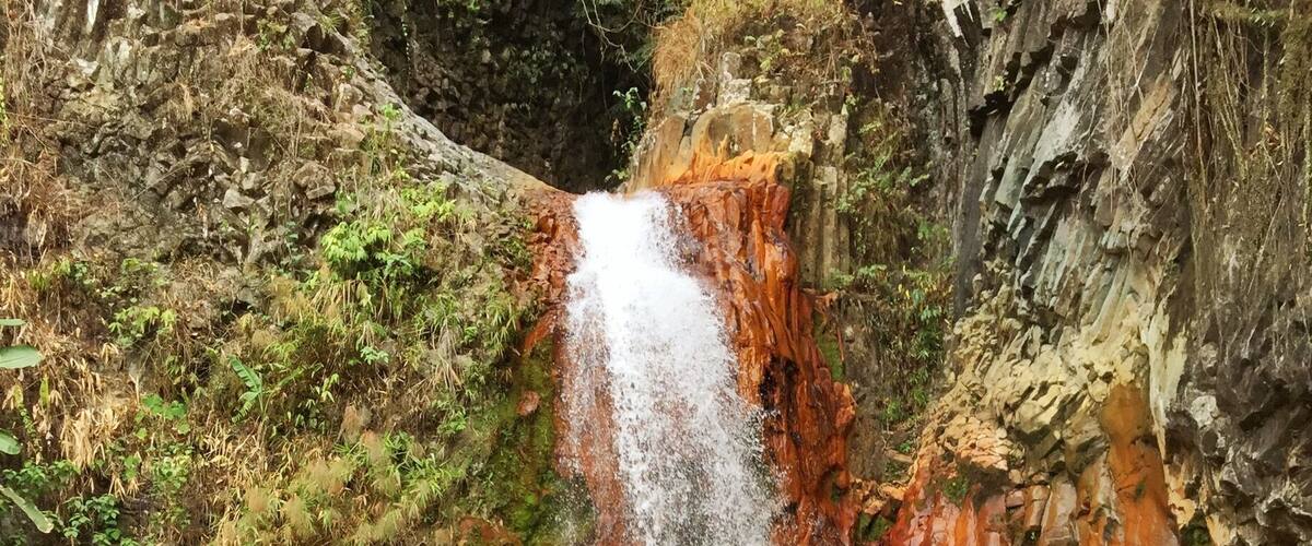 A waterfall cascading over reddish-brown rocks into a shallow stream with large boulders.