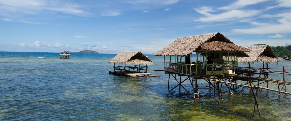 Overwater bungalows with thatched roofs on stilts in clear blue water.