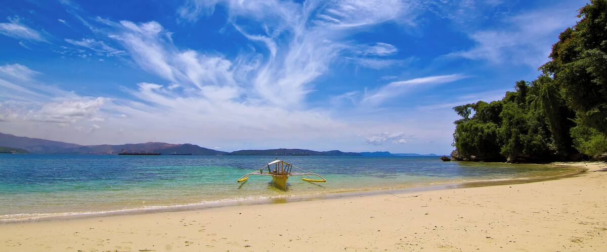 A beach with clear blue water, a boat, and a mountainous landscape in the background.