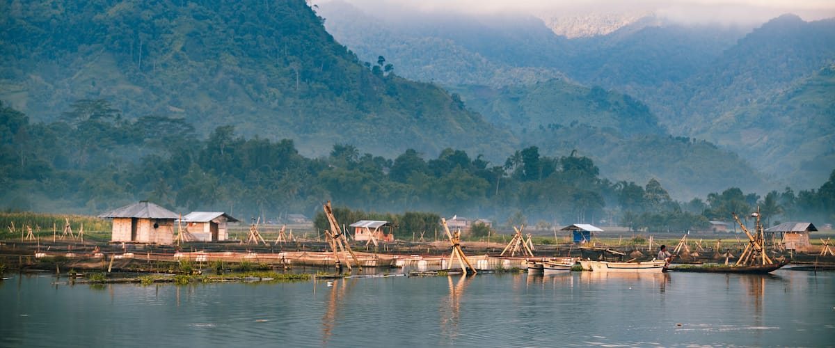 A calm river with traditional boats and wooden structures on the shore, surrounded by lush green hills and misty mountains.