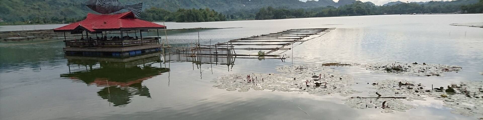A floating structure with a red roof, surrounded by calm water with lily pads and a distant mountain range.