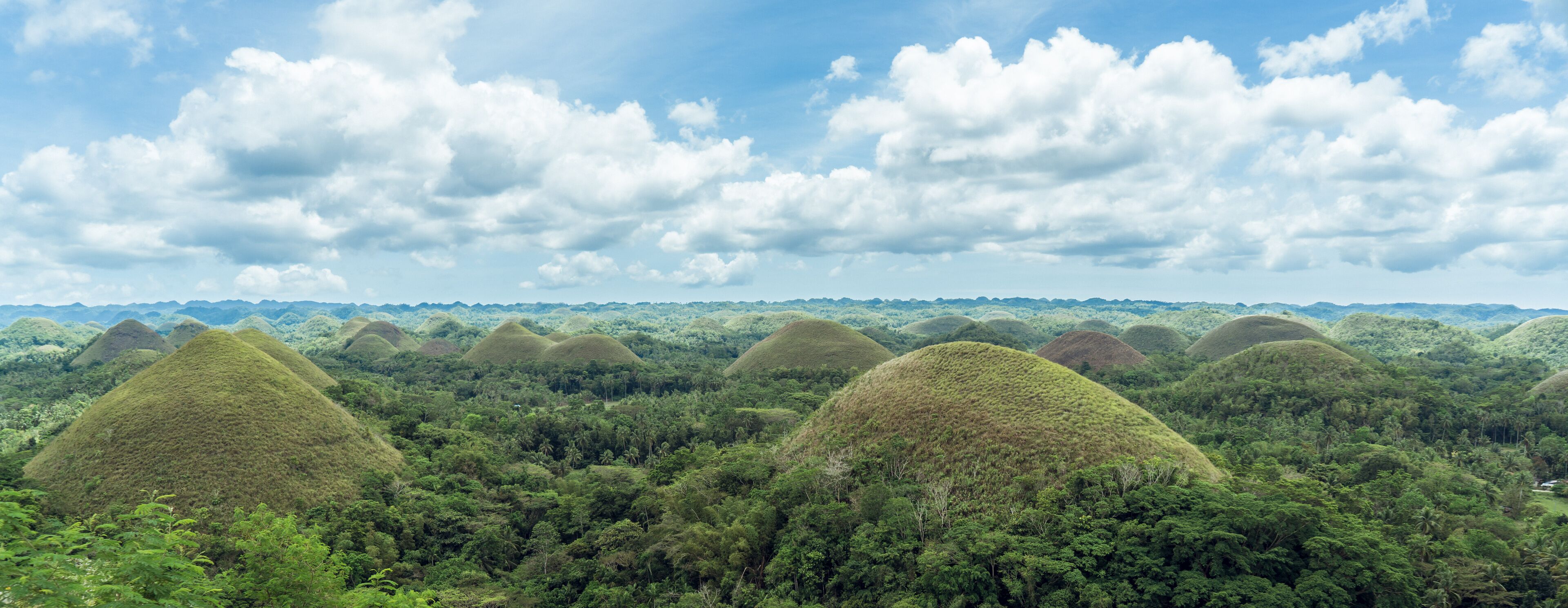 A landscape with numerous conical mounds surrounded by dense green forest under a partly cloudy sky.