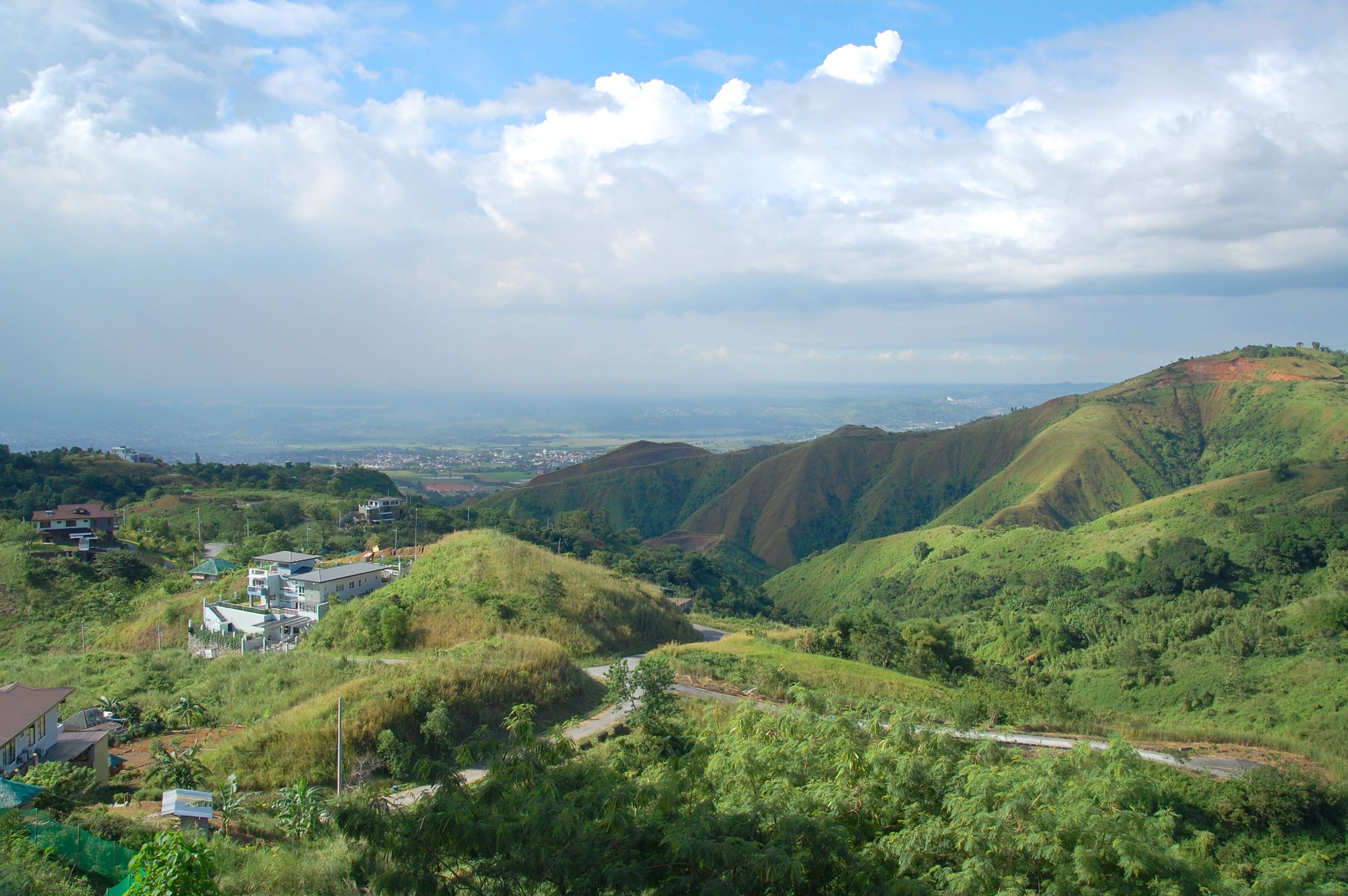 A valley with lush green hills, a winding road, and a distant cityscape under a partly cloudy sky.