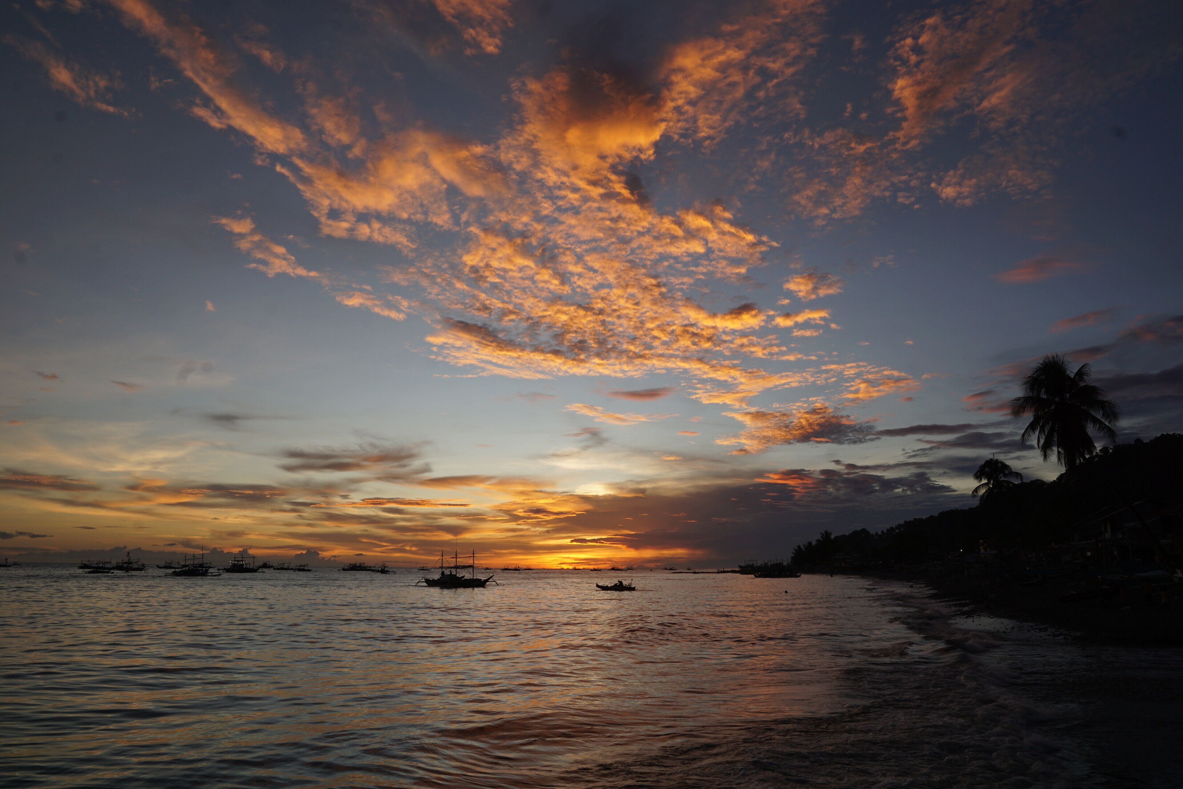 A sunset over a calm sea with boats and palm trees silhouetted against the sky.