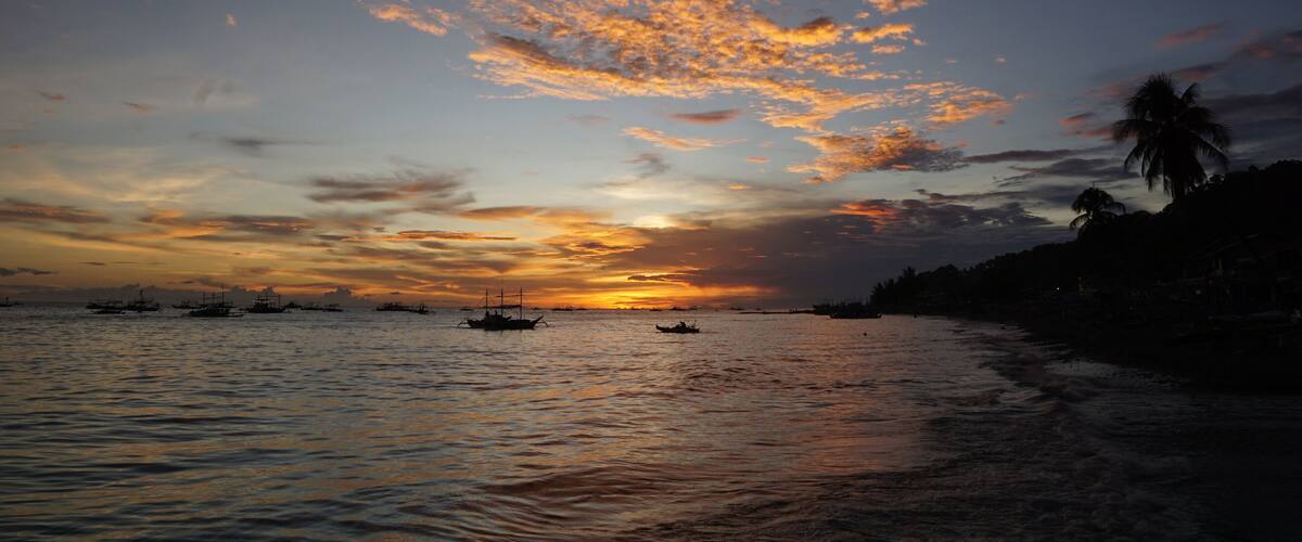 A sunset over a calm sea with boats and palm trees silhouetted against the sky.