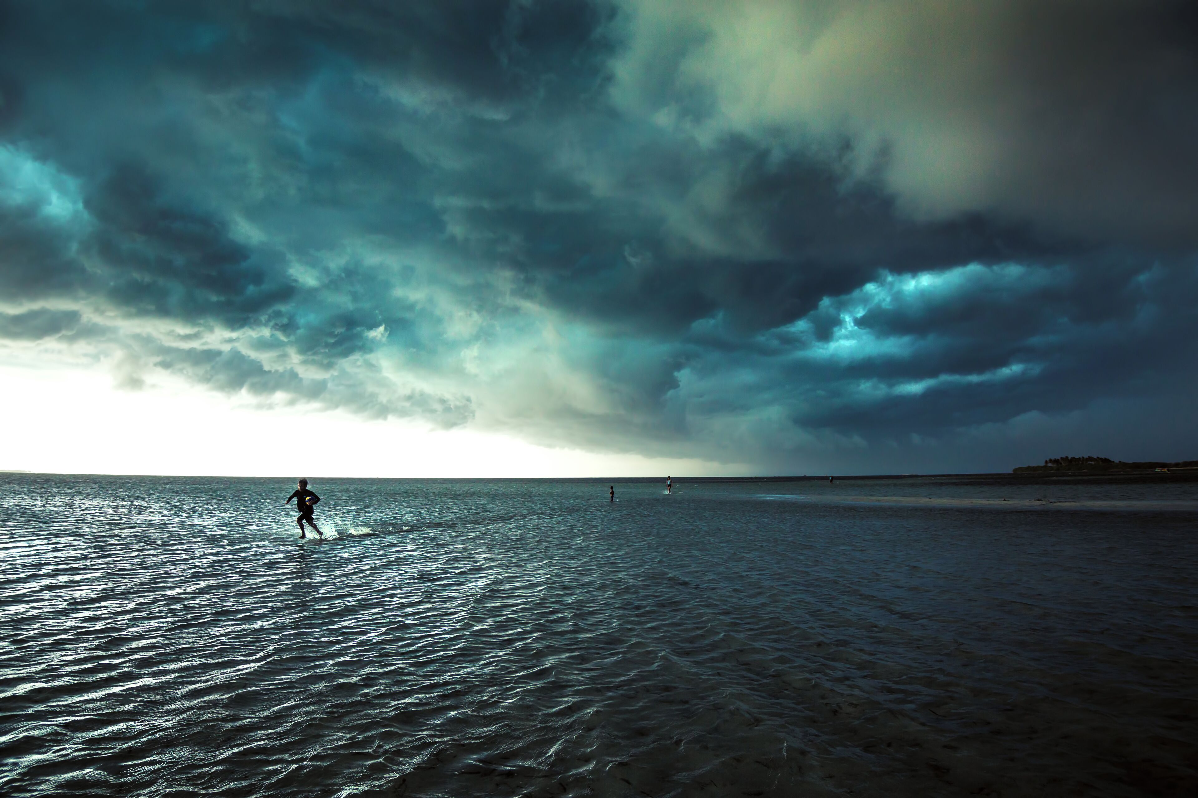 A beach with a person running in the water under a cloudy sky.