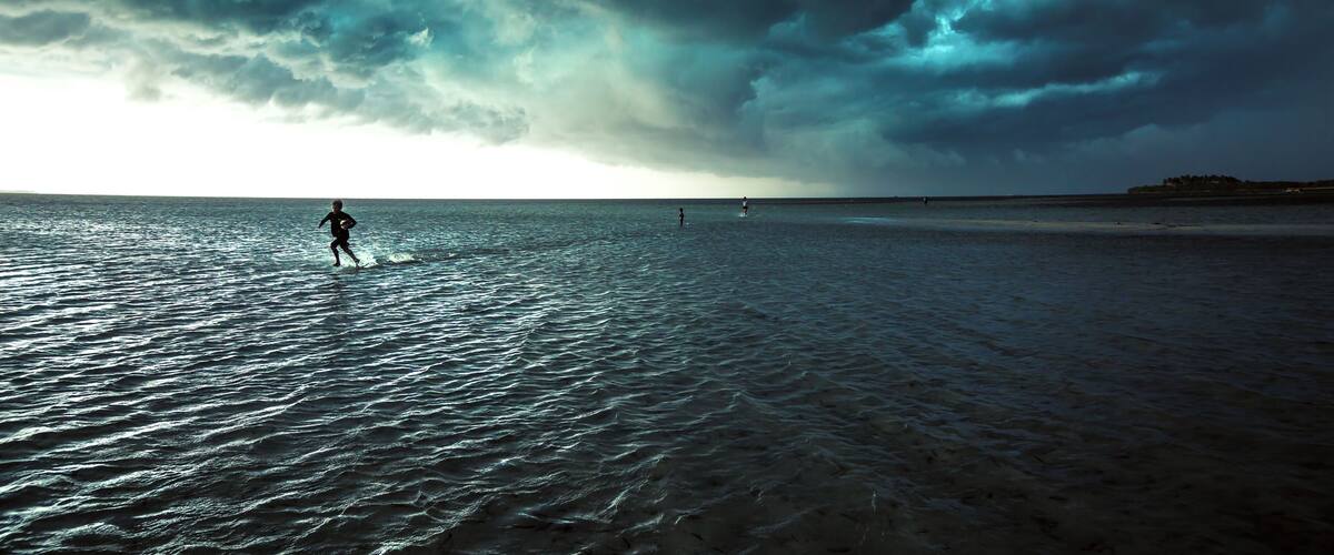A beach with a person running in the water under a cloudy sky.