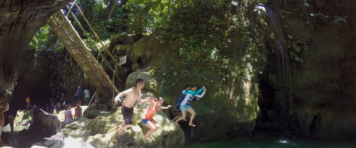 People jumping off a rocky cliff into a green pool.