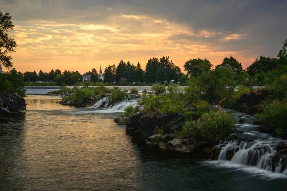 A river with waterfalls, surrounded by trees and a distant building.