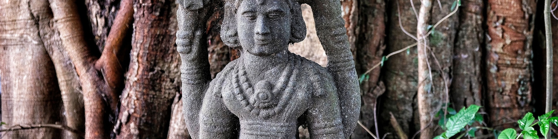 Estátua de pedra de uma figura sentada com um headdress, rodeada por raízes e folhagem de árvores.
