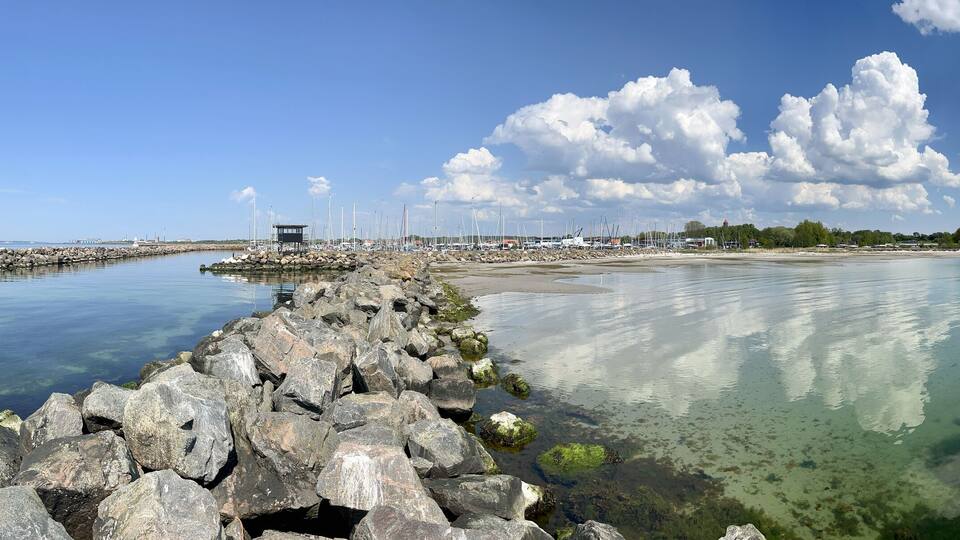 A calm waterfront with a rocky breakwater, clear water, and a distant shoreline with trees and buildings.