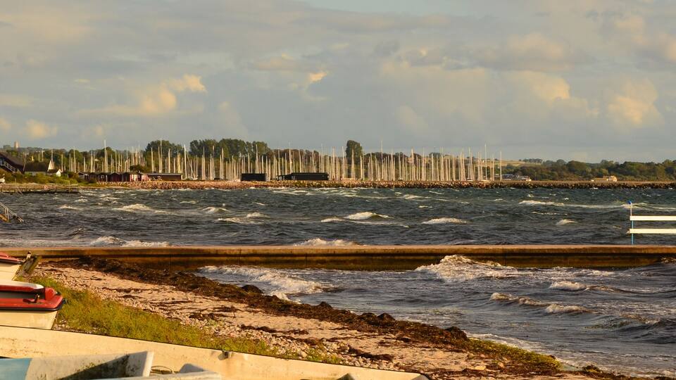 A coastal marina with boats docked, a breakwater, and a lighthouse in the distance.