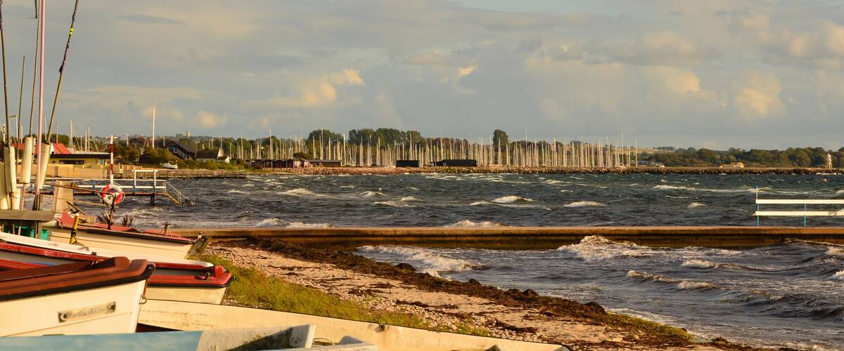 A coastal marina with boats docked, a breakwater, and a lighthouse in the distance.