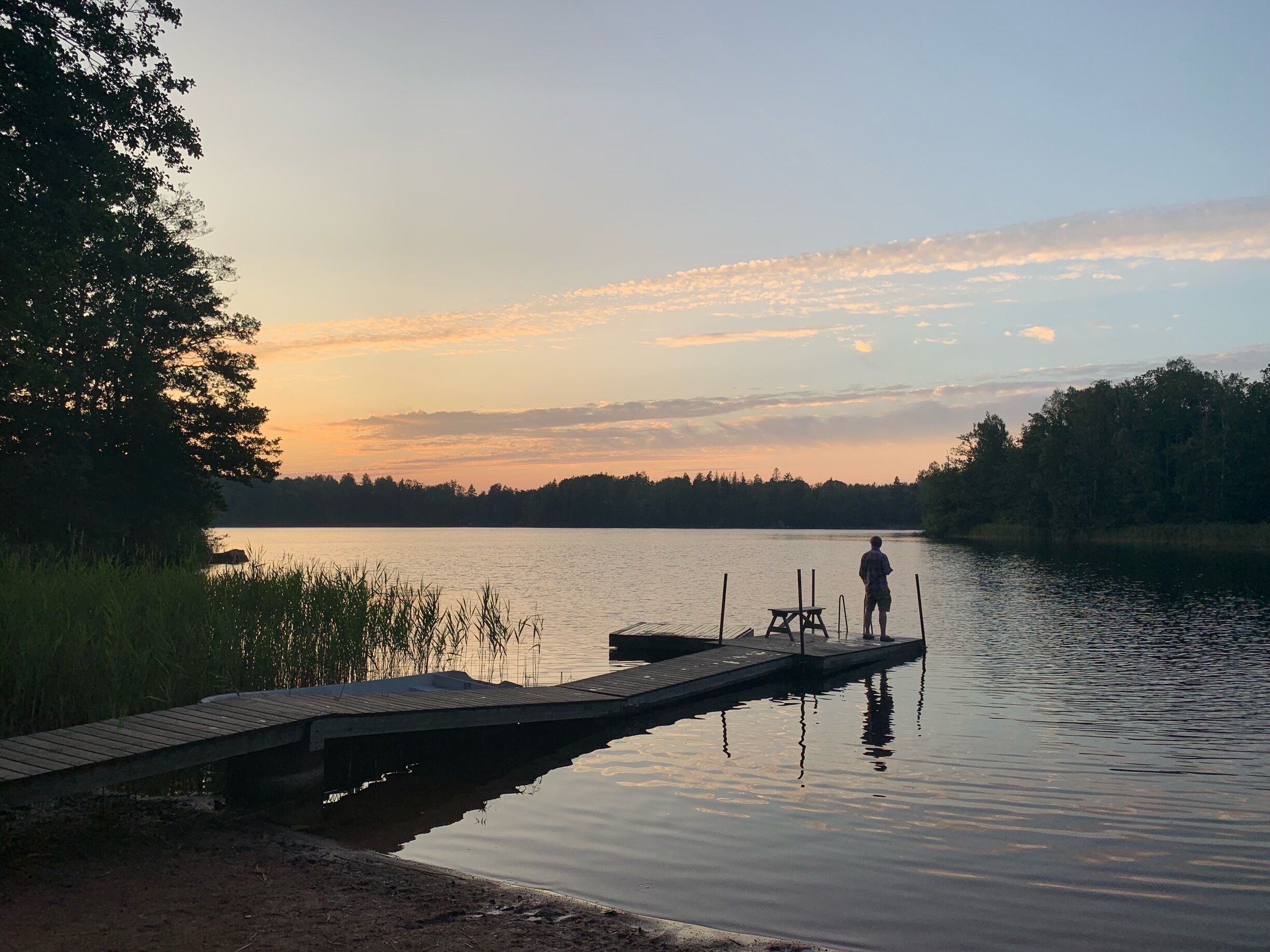 A wooden dock extending into a calm lake with a person standing at the end, surrounded by trees and a sunset sky.