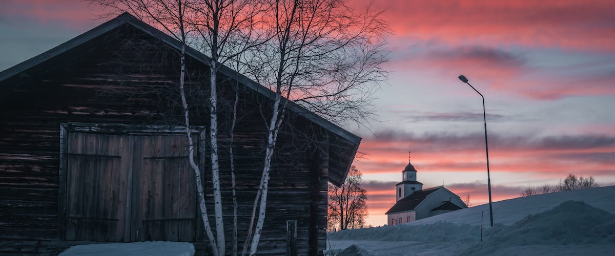 A wooden barn, a church, and a snowy landscape at sunset.
