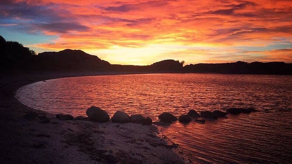A sunset over a beach with rocks in the foreground and hills in the background.