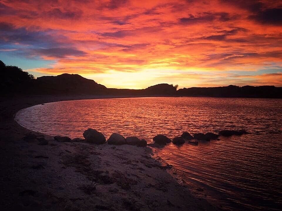 A sunset over a beach with rocks in the foreground and hills in the background.