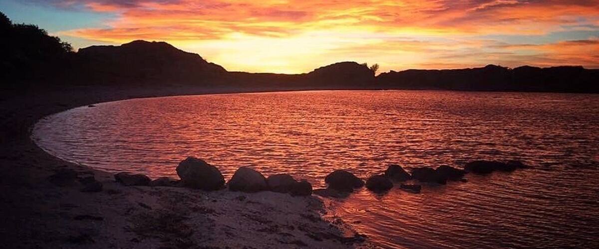 A sunset over a beach with rocks in the foreground and hills in the background.