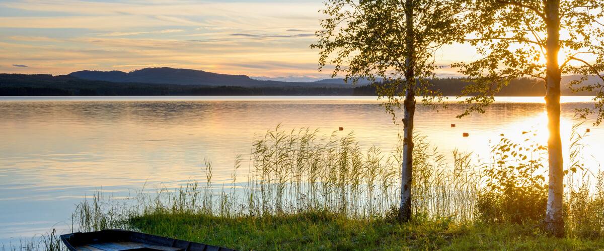 A wooden boat on calm water near a grassy shore with trees and a sunset in the background.