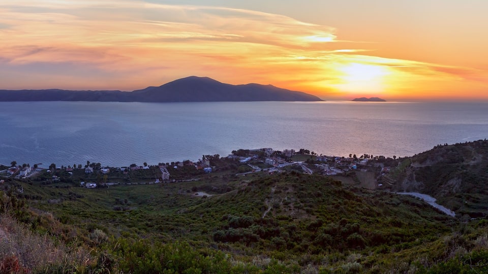 A coastal landscape with a sunset over the sea, hills, and a small town.
