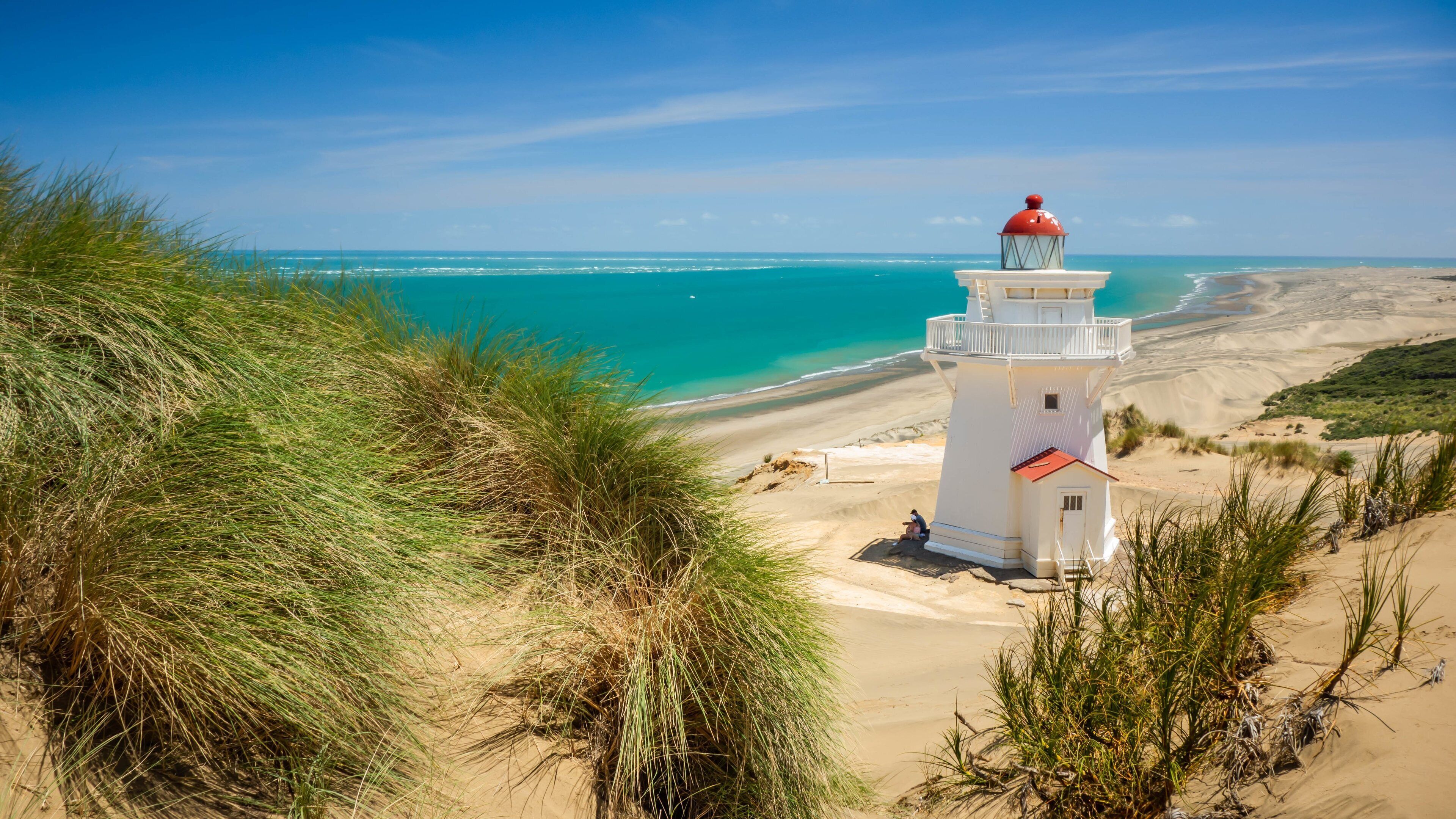 A white lighthouse with a red top, surrounded by sand dunes and coastal vegetation.