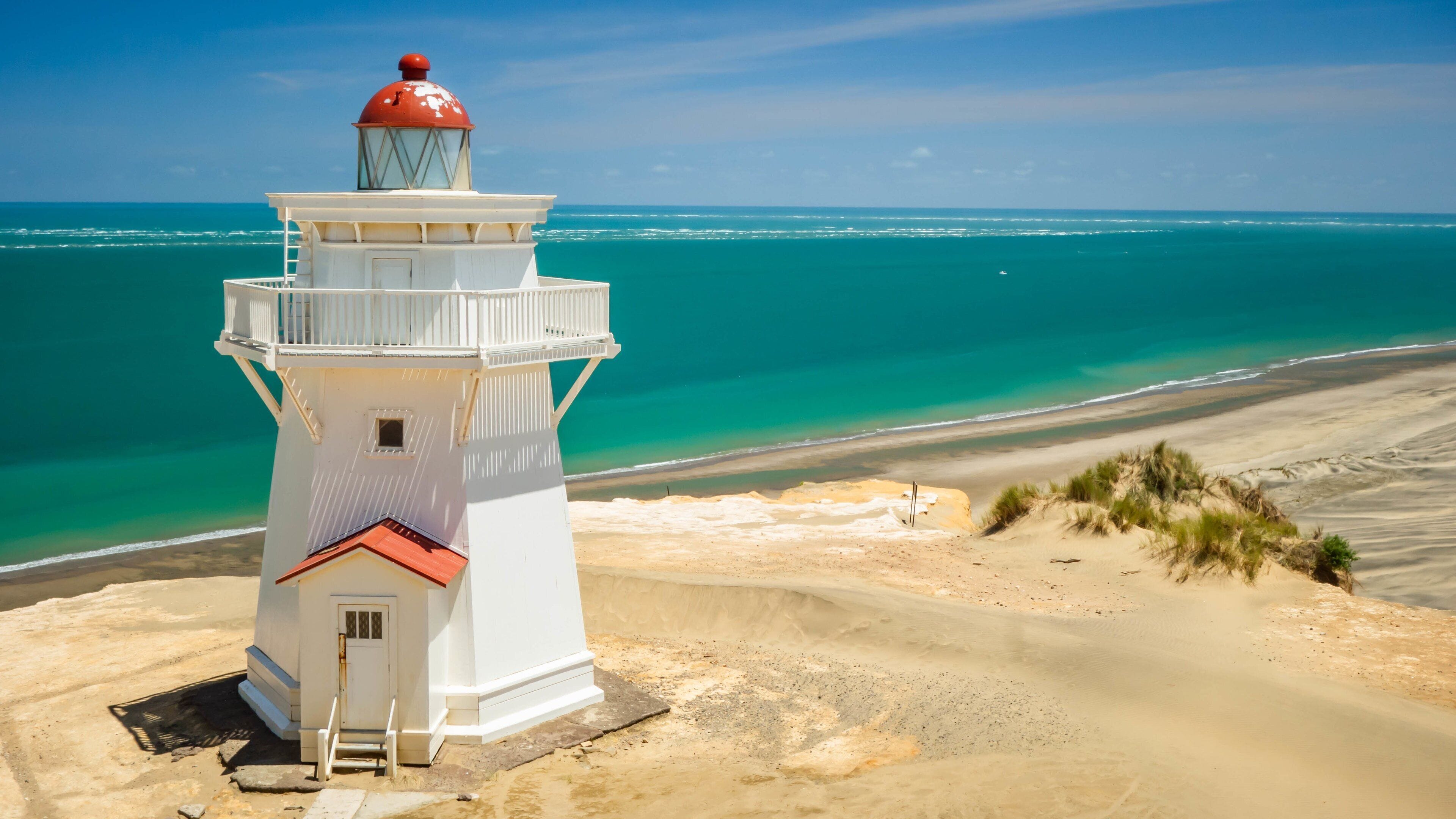 Un faro blanco con la parte superior roja, ubicado en una playa arenosa con aguas turquesas cristalinas al fondo.