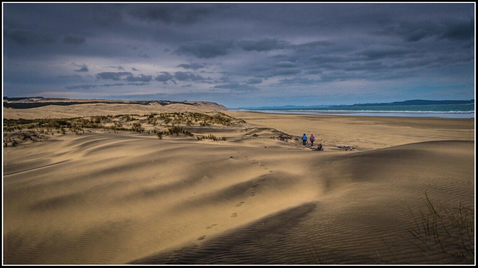 Un extenso paisaje desértico con dunas de arena, vegetación escasa y algunas personas caminando.