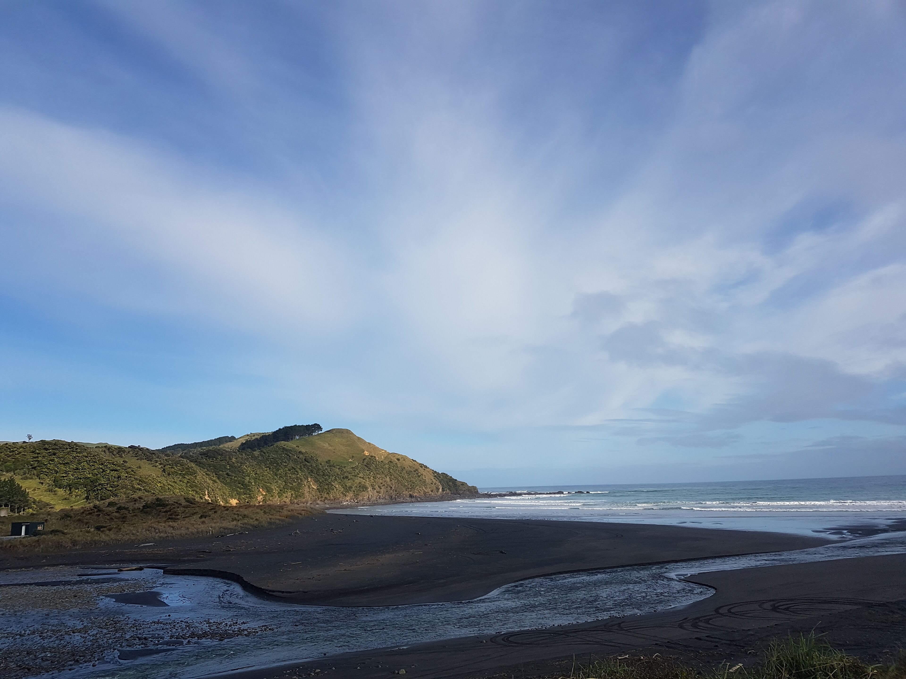 A beach with a dark sand shore, waves, and a hill with sparse vegetation.
