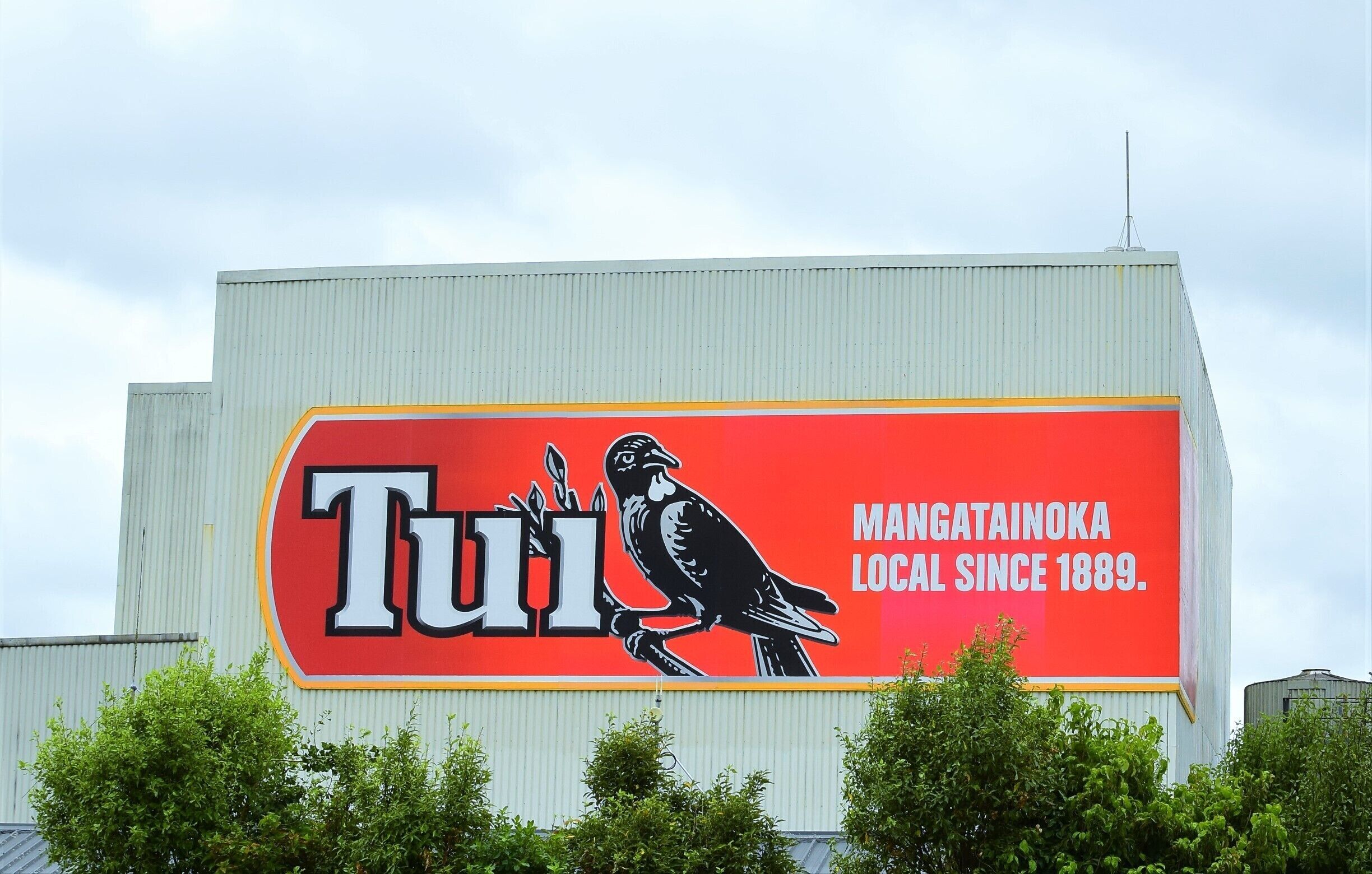 A large billboard with a red background and a black bird illustration, featuring the word 'Tui' and the text 'Mangatainoa Local since 1889.'
