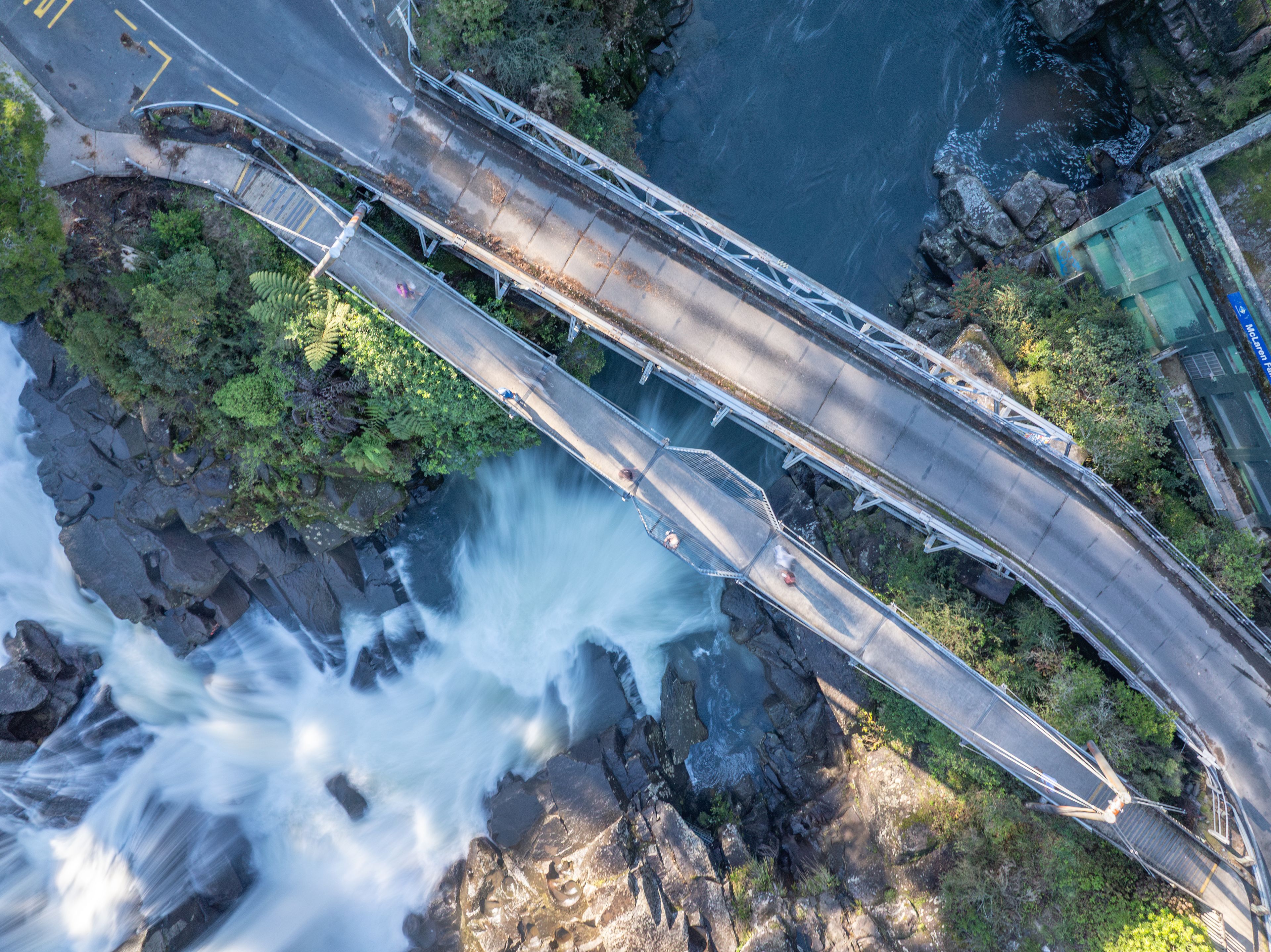 A bridge over a waterfall with surrounding greenery and a building in the background.