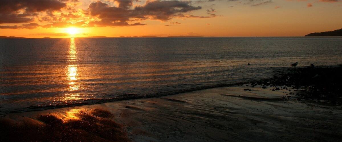 A sunset over a calm sea with a sandy beach and distant land.