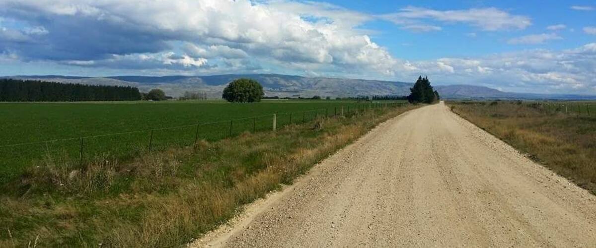 A gravel road in a rural area with fields, a fence, and distant mountains under a partly cloudy sky.
