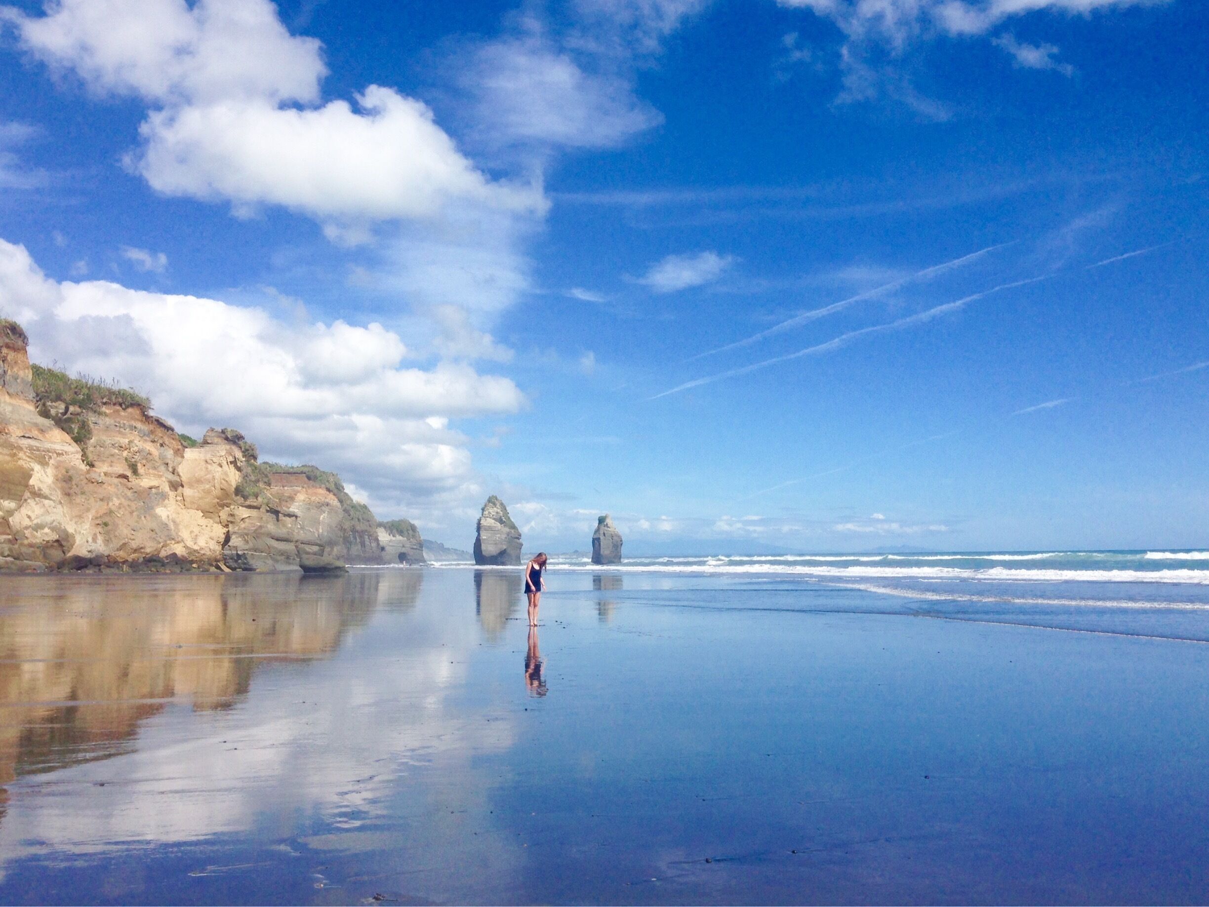 A beach with calm waters, a person standing, and rock formations in the distance.