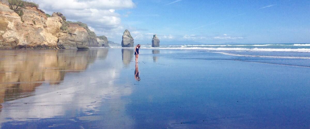 A beach with calm waters, a person standing, and rock formations in the distance.