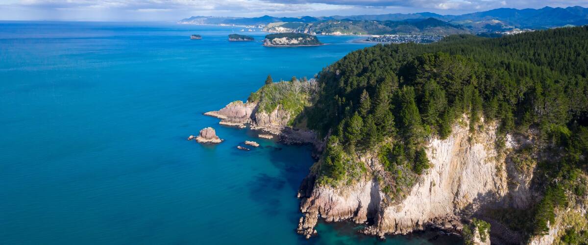 A coastal landscape with a forested cliff, clear blue sea, and distant mountains.