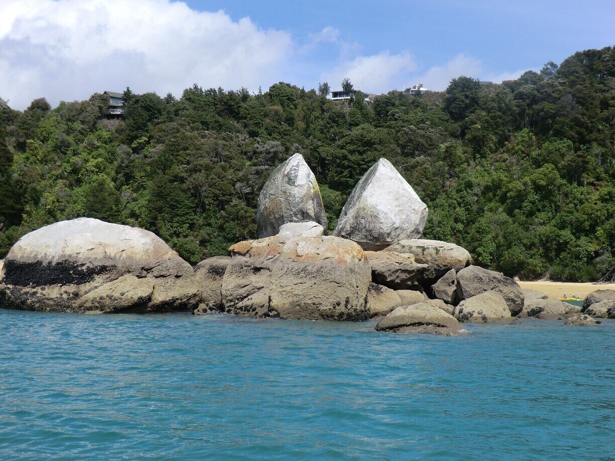 A coastal landscape with large boulders and a forested hill in the background.