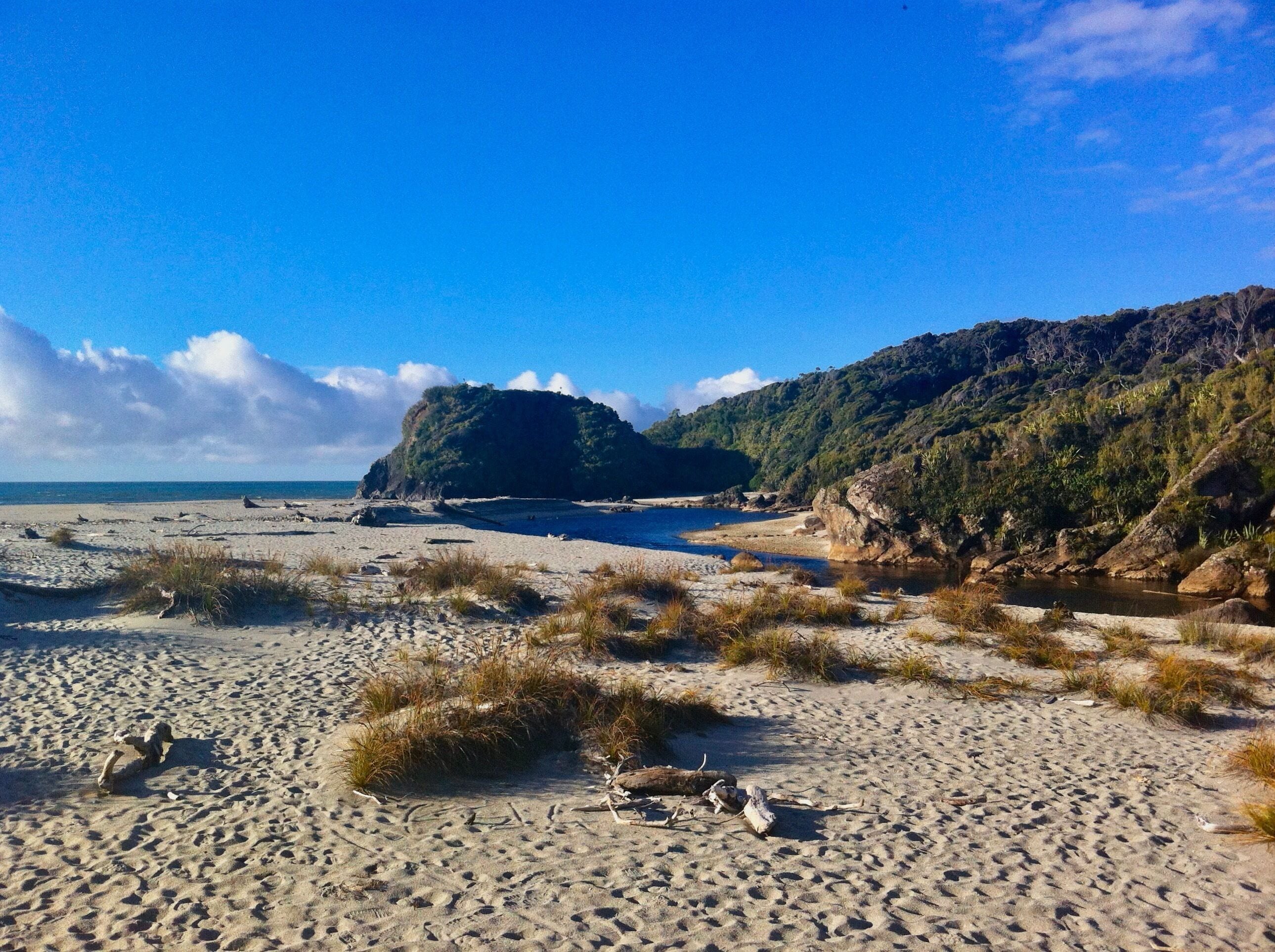 A sandy beach with scattered vegetation, a large rock formation, and a clear blue sky.