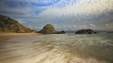 A beach with clear water, sandy shore, and rocky outcrops under a partly cloudy sky.