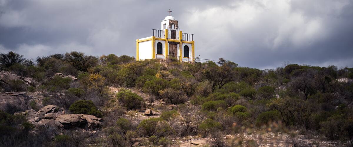 A white and yellow church on a hill with a cross on top, surrounded by shrubs and rocks.
