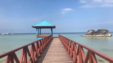 Un muelle de madera que conduce a un pabellón con techo azul, con vista a un mar tranquilo con aguas cristalinas y una pequeña isla con rocas al fondo.