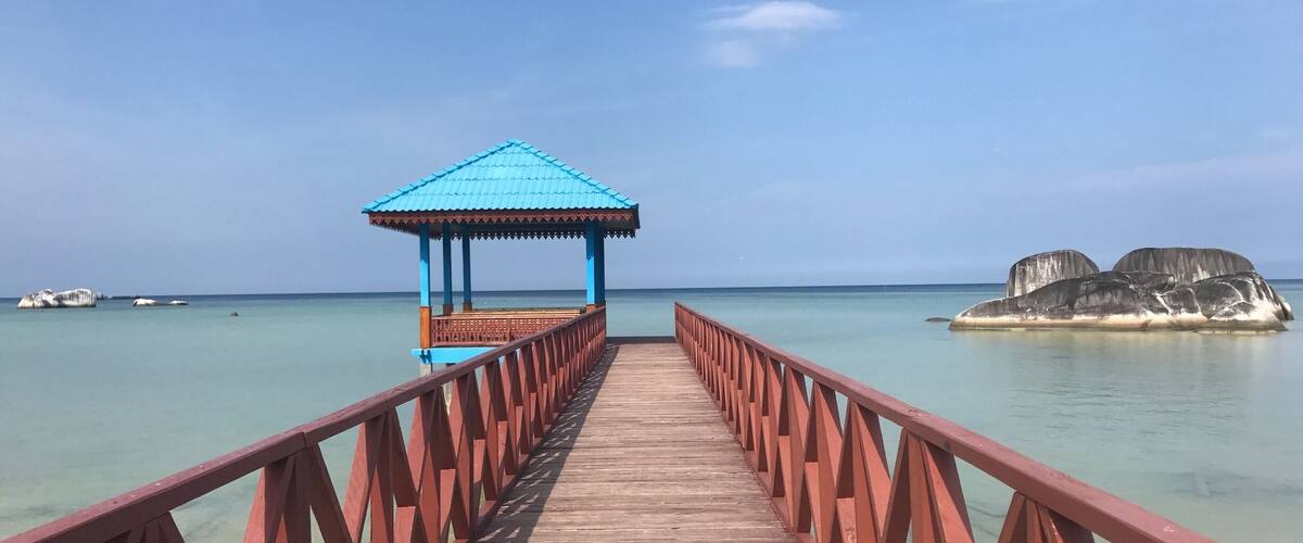 A wooden pier leading to a blue-roofed gazebo overlooking a calm sea with clear water and a small island with rocks in the distance.